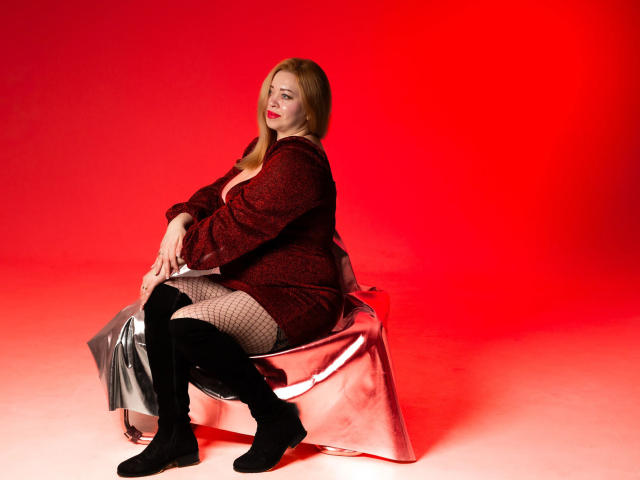 OliviaVerr sits on a shiny silver chair wearing a red patterned dress, fishnet stockings, and black knee-high boots, with red lighting highlighting her curvy ass and red lipstick.