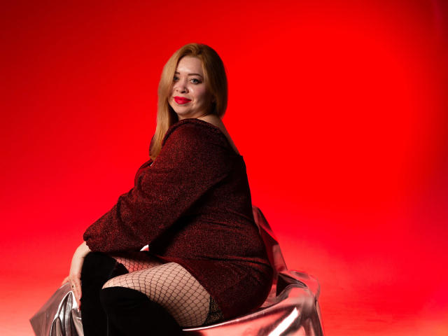 OliviaVerr sits on a silver-covered chair against a bright red background. She has long blonde hair and wears a sparkly red off-the-shoulder dress, fishnet stockings, and black knee-high boots. Her red lipstick matches the background, creating a bold contrast. She looks over her shoulder with a slight smile. The texture of the dress and the fishnet stockings adds depth to the image. The photograph uses strong, contrasting colors and focuses on her confident, relaxed pose.