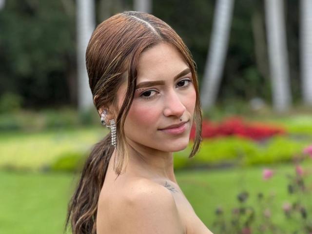 A portrait of AfroditaWild with light brown hair in a ponytail, looking over her shoulder. She has fair skin, brown eyes, and a small tattoo on her right shoulder. She's wearing clear, dangling earrings. Her face has a subtle blush and a slight smile. The background is blurred greenery with red and yellow flowers. The image has a natural, outdoor setting. The composition focuses on AfroditaWild's upper body and face.
