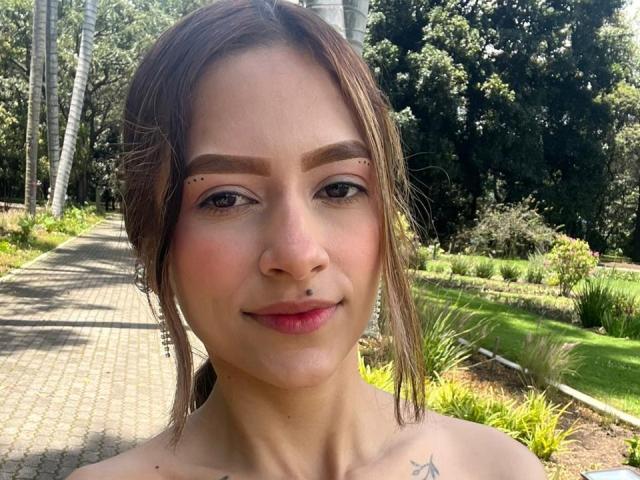 Close-up of AfroditaWild with light brown hair, small nose ring on the right, and eyebrow stud on the left. She has subtle makeup, pinkish blush, and a small star tattoo on her right collarbone. She's smiling slightly. Background shows a sunlit path, greenery, and trees. The image has natural light, clear focus on AfroditaWild's face, and a relaxed outdoor setting. Her hair is partially tied back.