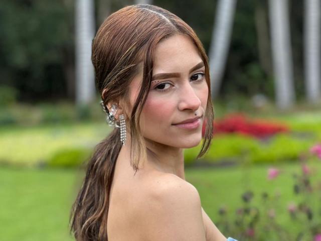 AfroditaWild with brown, wet-looking hair in a loose ponytail. She's facing slightly to the right, smiling subtly. She has fair skin, brown eyes, and is wearing large, sparkling earrings. Her bare shoulders are visible. The background is blurred with greenery and colorful flower beds. The image has a natural, outdoor feel with soft, even lighting. Her hair texture is smooth, and her makeup is minimal. The overall composition is balanced and focuses on AfroditaWild's upper body.