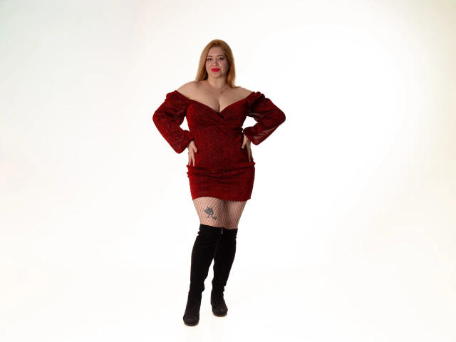 OliviaVerr stands with hands on hips, wearing a red off-shoulder dress, black fishnet stockings, and thigh-high boots, showing a tattoo on her right thigh.