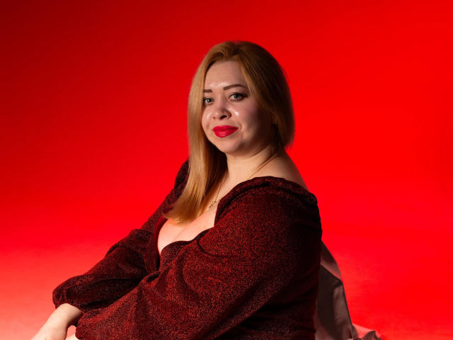 OliviaVerr with red lipstick, medium tits, and auburn hair, wearing a sparkly dark red dress, sitting against a bright red background. She's smiling slightly, looking over her shoulder.