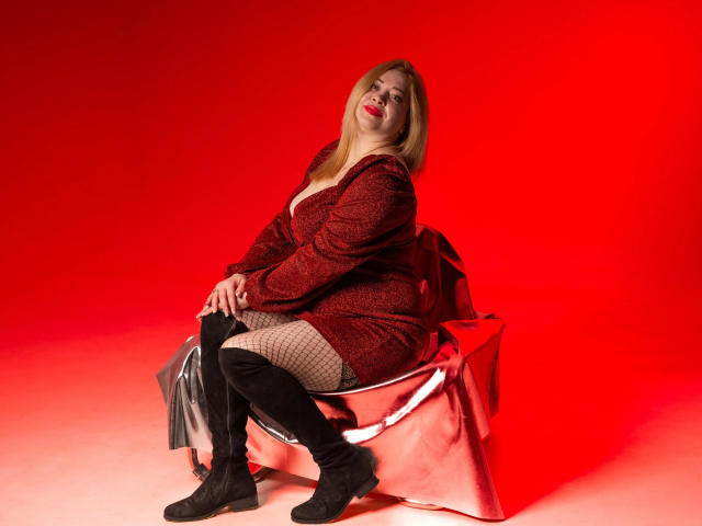 OliviaVerr sits on a shiny, metallic chair against a vivid red background. She wears a dark red, patterned dress with a plunging neckline, black fishnet stockings, and black knee-high boots. Her blonde hair is straight and falls past her shoulders. She looks slightly to the side with a relaxed, confident expression. The red light casts a warm glow over her, highlighting her fair skin and red lipstick. The metallic chair reflects the light, adding a modern, industrial feel to the composition.