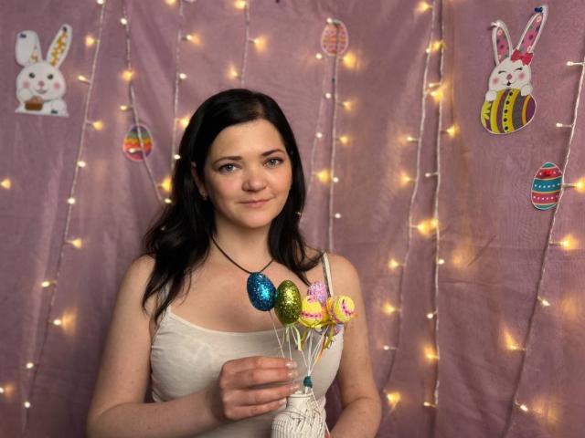 LauraFinn with dark brown hair, wearing a white tank top, holding colorful Easter egg lollipops, standing in front of pink fabric with fairy lights and bunny decorations.