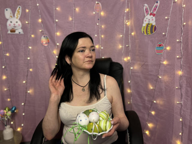 LauraFinn with black hair, pale skin, small tits, wearing a white tank top, holding green and white Easter eggs, sitting in a black chair, fairy lights in background.