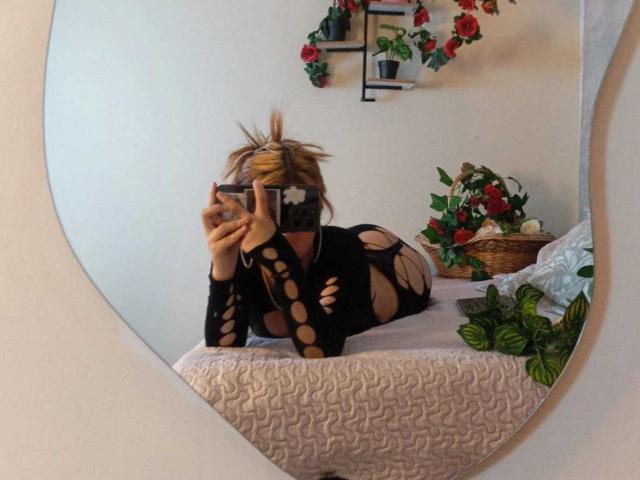 KatheLux lying on a bed, wearing a black, cut-out, fishnet bodysuit, with her blonde hair in a messy bun, holding a phone in front of her face.