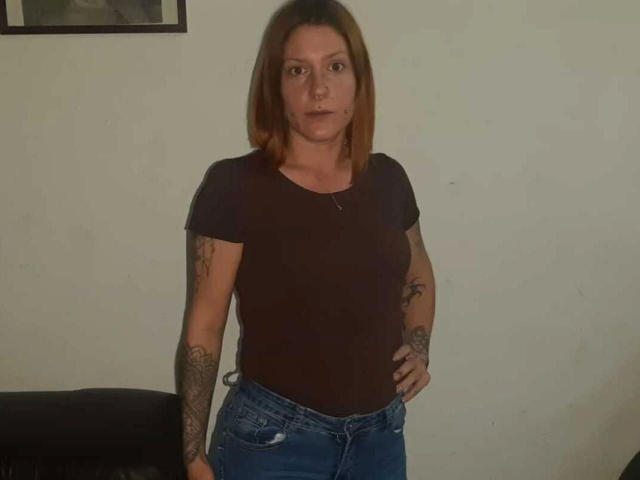 NinfaDeseo-hot has medium brown hair, a nose ring, tattoos on both arms, a black t-shirt, and blue jeans, standing with hands on hips, looking serious.