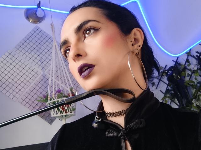 CandyParis with dark makeup, purple lipstick, hoop earrings, choker, black hoodie, and black leather flogger, looking up, blue neon lights in background, medium skin, black hair.