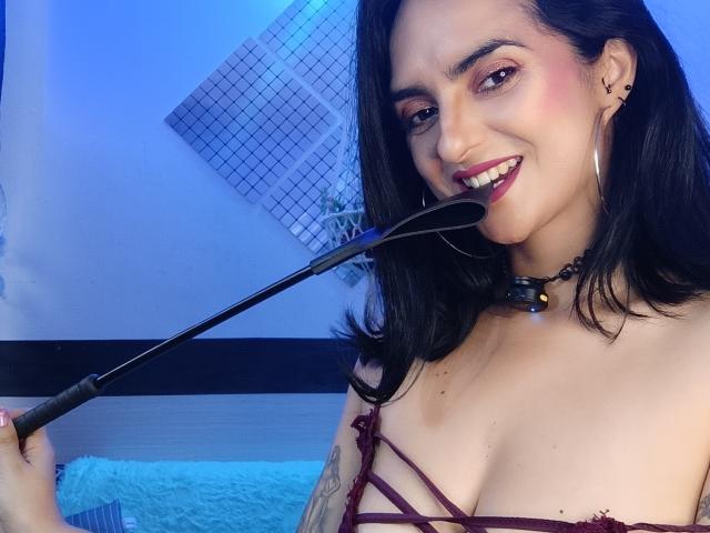 CandyParis, com cabelo preto, sorrindo, segurando um chicote preto, vestindo uma blusa vermelha com alças finas, um colar, brincos grandes e um piercing no nariz, mostrando seus seios. Iluminação azul..