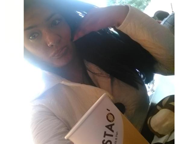 A photograph of ArianithaXox with long black hair, fair skin, and blue eyes, holding a Starbucks cup in a white hoodie. Her right hand touches her head. Bright sunlight creates a strong backlight, highlighting her face and hair. The cup has a white top with "STARBUCKS" in bold brown letters. Her expression is neutral. The image is slightly overexposed.