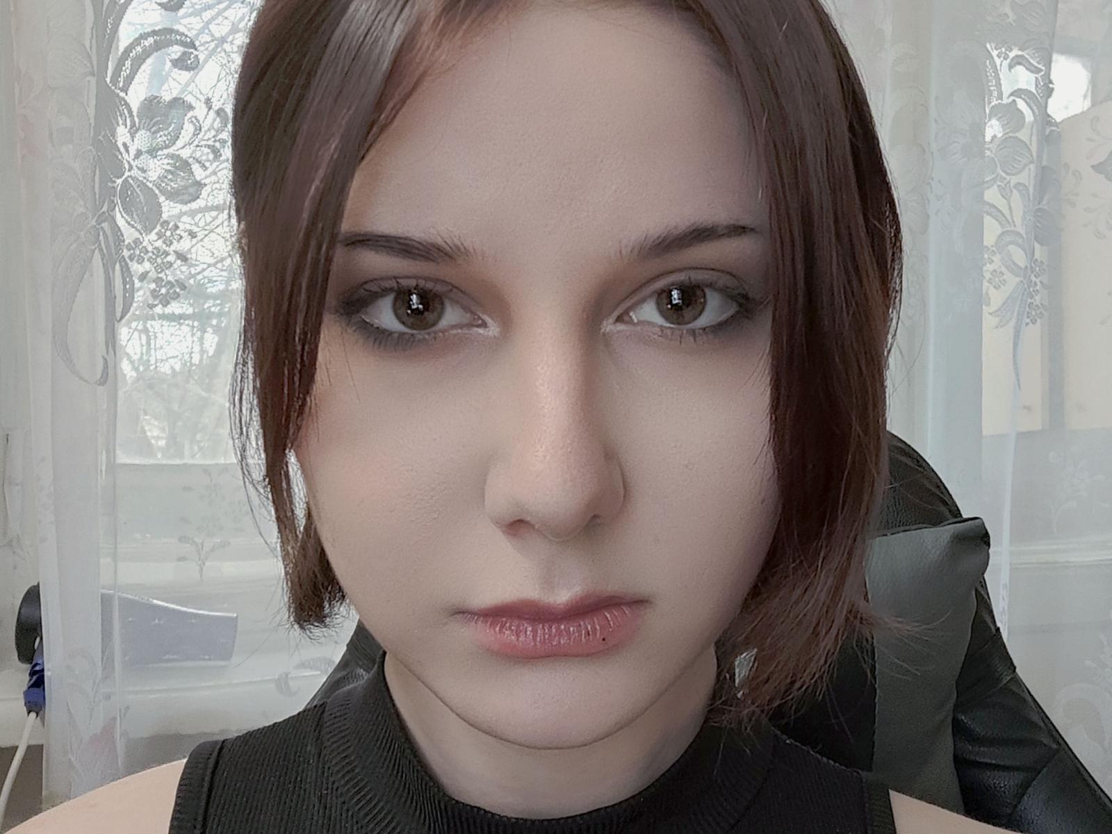 ImeldaBurckhard has pale skin, brown eyes, and short brown hair, wearing black, with a serious expression, and visible lips slightly parted, looking directly at the camera.