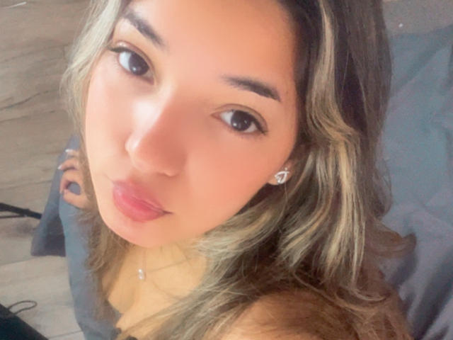 AmbarDunn with pouty pink lips, brown eyes, wavy blonde hair, and small earrings, looking up at the camera. She's topless, showing her tits. Her hands are on a blue surface.