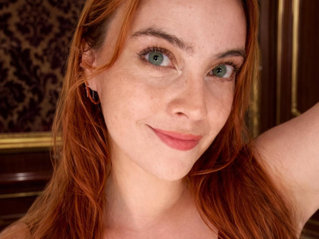 Close-up of Zireth, a fair-skinned woman with red hair and blue eyes. She has a slight smile, light freckles on her cheeks, and wears small, dark earrings. Her hair is loose, and she raises her right arm. The background features a dark, patterned texture and wooden elements. The lighting is even, highlighting her facial features. The image focuses on her facial expression and eye color.