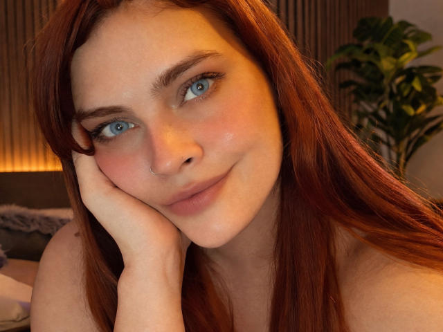 Close-up of Zireth with fair skin and red hair, head resting on hand. Zireth has blue eyes, subtle freckles, and a small nose ring on the right side. Zireth's lips are slightly parted in a subtle smile. The hair is straight and falls over shoulders. The background is blurred, with a potted plant on the right. Warm lighting highlights Zireth's face. The image has a natural, relaxed feel. Zireth's gaze is direct and confident.