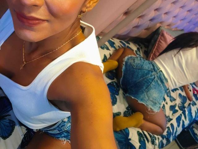 HannaPretty with tan skin, small tits, wearing a white ribbed tank and frayed denim shorts, yellow socks, and gold necklace, kneeling on a bed with blue floral pattern.