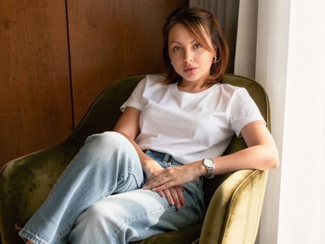 AnitaLevantaa sits in a green velvet chair, wearing a white tee, blue jeans, and a silver watch, legs crossed, hands on her lap, looking straight at the camera.
