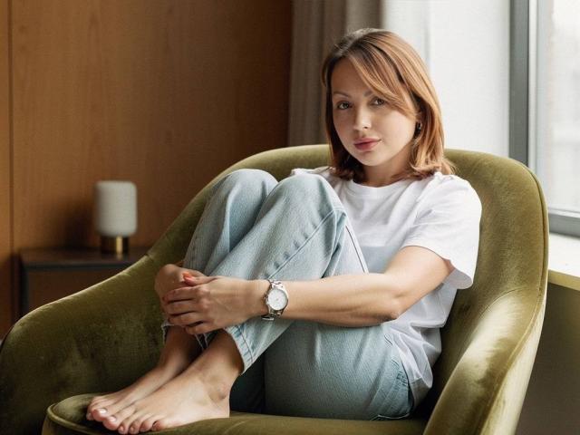 AnitaLevantaa is sitting in a green velvet chair, legs crossed, wearing a white tee and blue jeans, barefoot, with a silver watch, looking calm and relaxed.