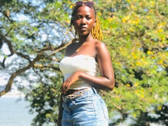 SexyBlack-hot with dark skin, yellow braids, wearing a white strapless top and blue jeans, standing outdoors, sunglasses on head, hand on belt, sunny day, green tree background.