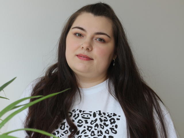 A portrait of MileyMiln with long, straight brown hair and fair skin. She wears a white t-shirt with a black leopard print design. MileyMiln has subtle makeup with pink lipstick and small earrings. Her eyebrows are dark and slightly arched. She looks directly at the camera with a slight, calm smile. A green plant leaf is in the lower left corner. The background is plain and light.