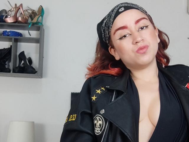 BelindaHedone has a light skin tone and red-dyed hair, wearing a black bandana and a black leather jacket with yellow stars and a patch on the left sleeve. She has a deep V-neck black top showing cleavage. Her makeup includes bold red eyebrows and winged eyeliner. She is making a pouty expression. The background has a shelf with high-heeled shoes in various colors and a small white lamp. The image is a photograph.
