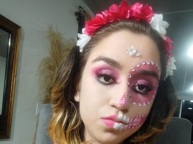 BelindaHedone's face is covered in pink and white makeup with glitter, pink lipstick, and a pink flower headband. She has brown hair, pink eyeshadow, and a serious expression.