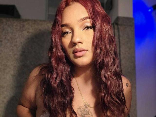 Valerie Wet tiene el pelo rojo ondulado, labios carnosos y un tatuaje en el pecho. Lleva un collar y tiene una ligera expresión de ceño fruncido. Sus senos son visibles..