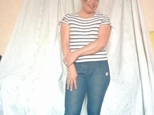Rahel69, standing with hands crossed, wearing a striped shirt and blue jeans, has a smile, small tits, and a ring on her right hand, against a white curtain.