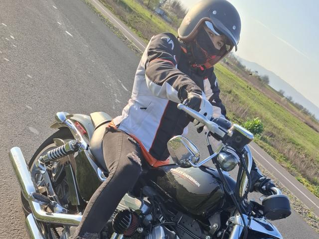 FrancaiseAnnaDom rides a green and silver motorcycle on a sunny road. Wearing a black helmet, reflective visor, and black and white jacket with red accents, FrancaiseAnnaDom grips the handlebars. The motorcycle's chrome details and large rear exhaust are visible. FrancaiseAnnaDom's black pants and gloves are in contrast with the motorcycle's metallic parts. The road and green field in the background are blurred. The image has a slight downward angle, emphasizing the motorcycle and rider.