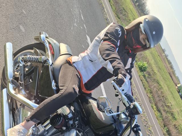 FrancaiseAnnaDom riding a black motorcycle with a chrome bar, wearing a helmet, black and white jacket, torn jeans, and gloves, on a sunny road.