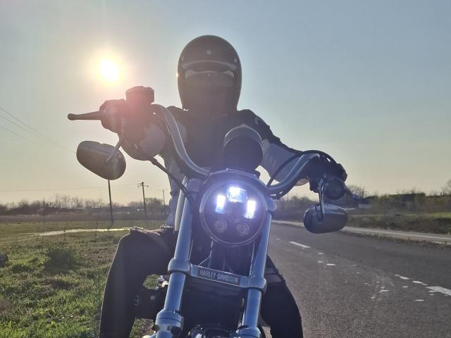 FrancaiseAnnaDom on a motorcycle, wearing a black helmet and gloves, with sun in the background, holding handlebars, front headlight on, and road in the foreground.
