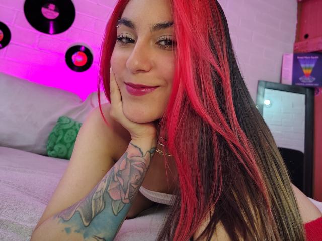 AmyStreetwise69 with red and black hair, smiling, resting head on hand, showing tattooed arm, pink lipstick, and white tank top, in a cozy room with purple and pink lights.