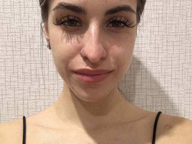 Close-up of DinorahYanke. She has light skin, brown eyes with long, dark eyelashes, and a small nose ring on her right nostril. Her lips are slightly parted in a subtle smile. She wears a black spaghetti strap top. Her brown hair is pulled back. The texture of the background is a light, patterned fabric. The image is centered, focusing on her face and upper shoulders. The lighting is even, highlighting her facial features. Her eyebrows are thick and dark, contrasting with her lighter skin.