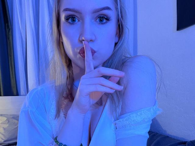 JanellLindinha with blue-tinged skin, blonde hair, and blue eyes, sucking her finger, wearing a white off-shoulder top, exposing her tits. She's making a "shh" gesture.