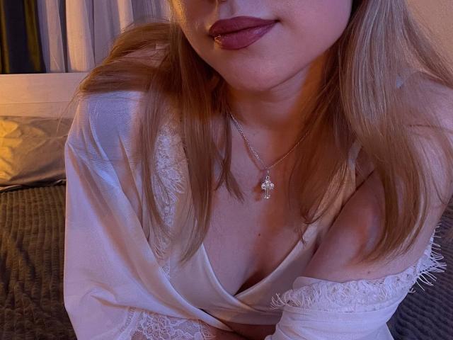 JanellLindinha vestindo um roupão branco com detalhes em renda, ligeiramente aberto para revelar seios pequenos, com um colar dourado e batom rosa, cabelo loiro e um sorriso discreto, em uma luz roxa suave..