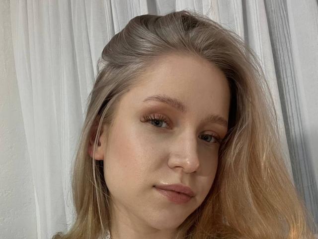 Close-up portrait of JanellLindinha with light blonde hair, slightly wavy, and fair skin. She has subtle, natural makeup with light pink lips and minimal eyeliner. Her green eyes look directly at the camera. The texture of her hair is smooth with some loose strands. She has a small, slightly upturned nose and a gentle expression. The background consists of white, slightly wrinkled curtains. The lighting is soft, highlighting her facial features. The overall composition is simple and focused on her face.