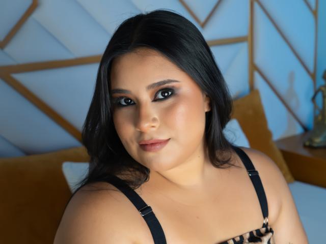 Close-up photo of SimonaAntury with medium skin tone, black hair, and dark brown eyes. She has a slightly rounded face with subtle makeup, including pinkish lipstick and dark eyeliner. She wears a black strap bra with a leopard print pattern. The background has a geometric, light blue and gold pattern. SimonaAntury's expression is neutral, with a slight, closed-mouth smile. The image has a soft, even light. The focus is on her upper body and face.