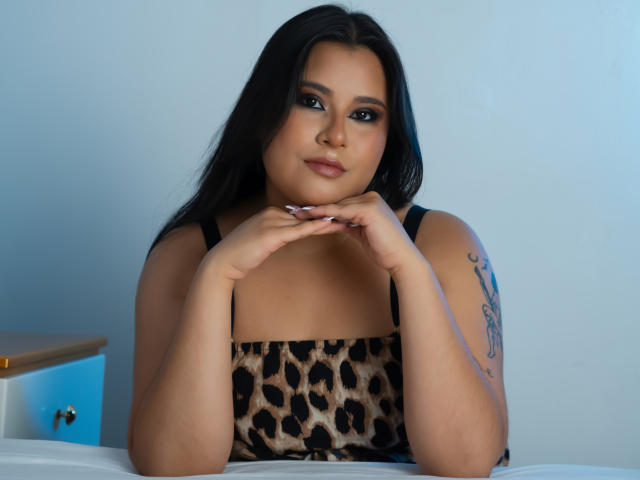 SimonaAntury has long black hair and dark eyes, wearing a leopard-print dress with black straps. She's sitting with hands gently touching her chin, looking slightly to the side. She has a tattoo on her right arm, featuring cursive text. The lighting is soft, highlighting her smooth, tan skin. The image has a blue tint, adding a cool atmosphere. She's centered, with a white bed and a wooden nightstand in the background. The overall composition is balanced and focused on SimonaAntury's relaxed yet confident expression.