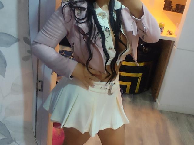 Diiana with long black hair, pink jacket, white shirt, and white pleated skirt, standing, showing her ass, hand on hip, in a room with wooden floor and yellow bag.
