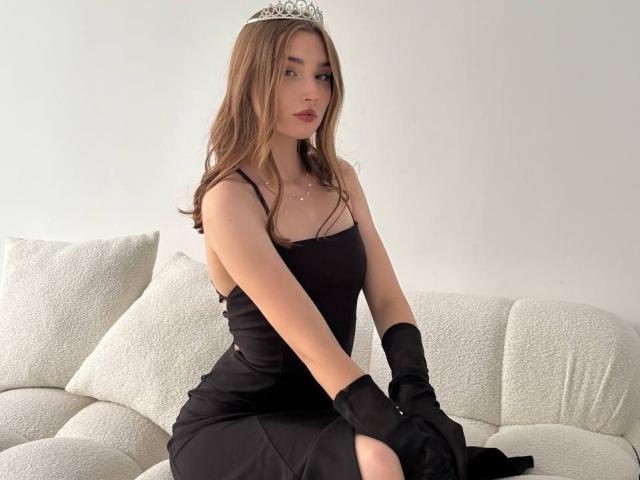 KandraPowlen, with brown hair, wearing a black dress, black gloves, and a silver tiara, sits on a white couch, showing her tits and looking serious.