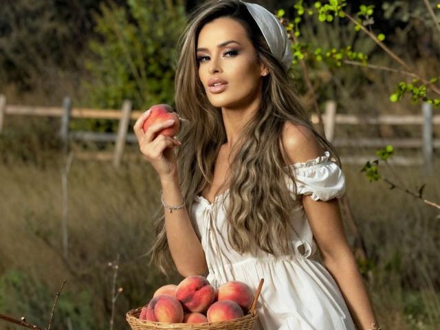 AshyQuinn con lunghi capelli castani ondulati, vestito bianco con spalline scoperte, che tiene una pesca, in piedi accanto a un cesto di pesche, illuminata dal sole, con foglie verdi sullo sfondo, che appare sensuale..