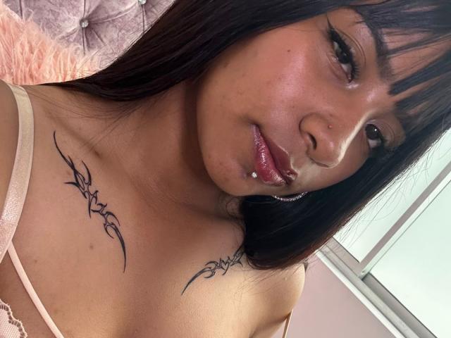 NahomyNoir has dark brown hair, full pink glossy lips with a piercing, and black tribal tattoos on her tits. She's wearing a beige lace top, looking at the camera.