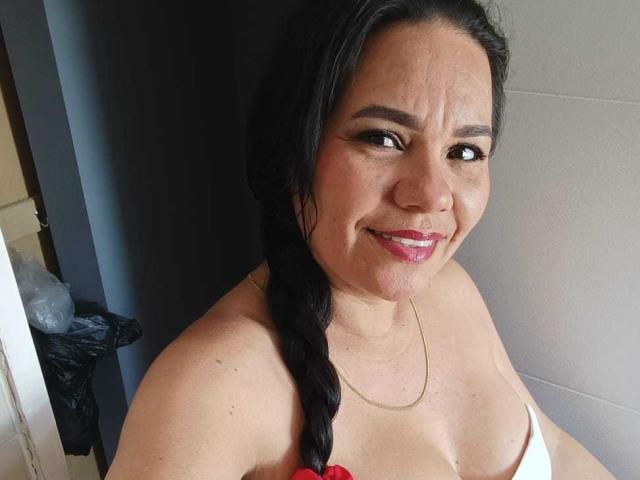 A photograph of KinkyMaturee, a middle-aged woman with tan skin and dark, straight hair in a single braid. She has thick eyebrows, black eyes, and is smiling with pink lipstick. She wears a white strapless top with a red floral design and a thin gold necklace. The background is a dark gray wall with a black trash can and cardboard box partially visible on the left. The image has a casual, slightly cluttered feel.