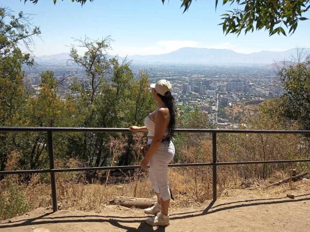KinkyMaturee stands on a dirt path, facing a city below. She's in a white strapless dress, white sneakers, and a white cap. Her long black hair is down. She leans on a metal railing with her right hand. The city's buildings are spread out, surrounded by green and brown foliage. Mountains are in the distant background. The sky is clear and blue. Her shadow is on the ground. She's on the right side of the image.