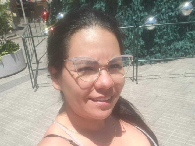 Close-up photo of KinkyMaturee, a woman with light brown skin and dark brown hair, wearing transparent glasses and a light pink top. She's smiling with a slight squint from sunlight. Her face is centered, showing a relaxed expression. She has a small nose and full lips. The background features a green, textured wall and string lights, with a potted plant on the left. The image has a bright, natural light. KinkyMaturee's glasses have a thin frame, and she's wearing a simple necklace.