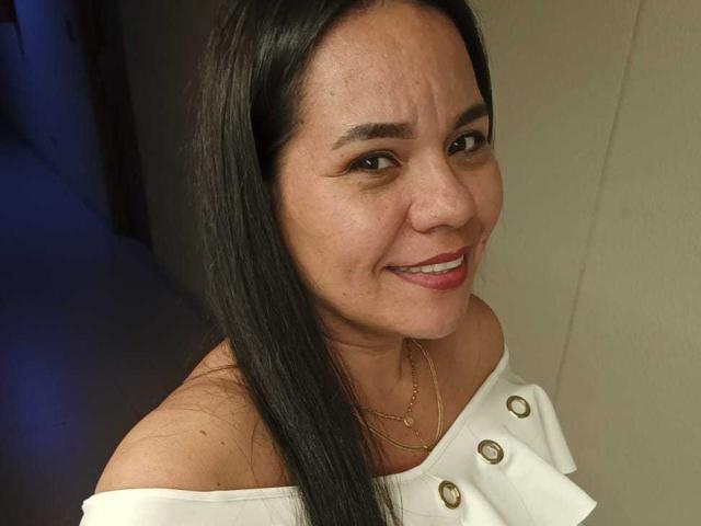 Close-up photo of KinkyMaturee, a smiling Asian woman with straight black hair, medium skin tone, and small freckles on her face. She's wearing an off-shoulder white top with gold buttons and a thin gold necklace. Her expression is friendly and slightly mischievous. The lighting is soft, highlighting her facial features and the texture of her top. Her hair is parted in the middle, and she's looking up at the camera. The background is blurred, with a dark hallway on the left.