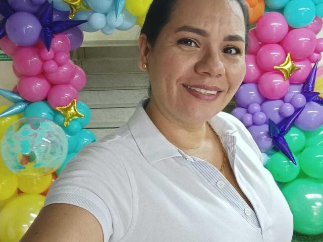 KinkyMaturee with dark hair, smiling, wearing a white polo shirt, standing in front of a colorful balloon arch with stars and a clear balloon. Boobs are not visible.