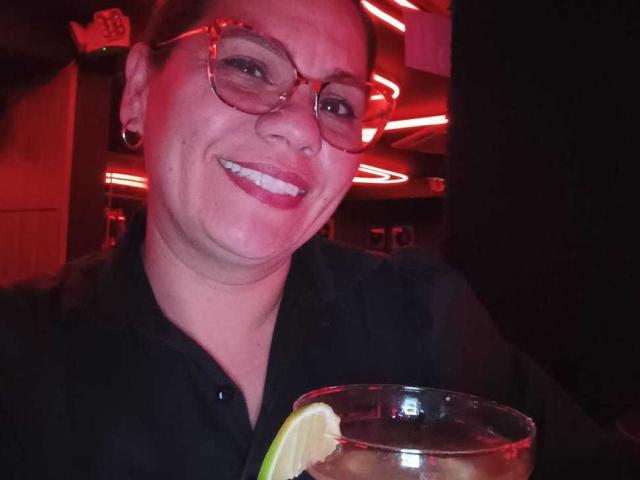 KinkyMaturee has a light skin tone, short brown hair, and wears glasses with a slight reflection. She's smiling with visible teeth, showing a bit of her upper lip. KinkyMaturee is dressed in a black shirt. She holds a clear glass with a lime wedge at the rim, filled with a dark liquid. The red lighting in the background creates a warm, almost neon glow on her face. KinkyMaturee has small black earrings. The image is slightly blurred, emphasizing the red light and her smile.