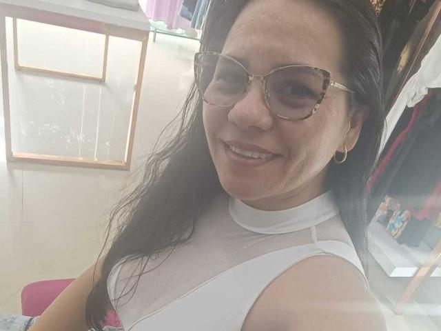 Close-up of KinkyMaturee with long dark hair, wearing gold-rimmed glasses, and a white sheer top. She has a fair complexion, a slight smile, and visible teeth. She's wearing a gold hoop earring in her left ear. The background has a glass shelf with clothes and a pink chair. The lighting is soft, highlighting her facial features and the texture of her top. The image has a casual, slightly blurry quality.