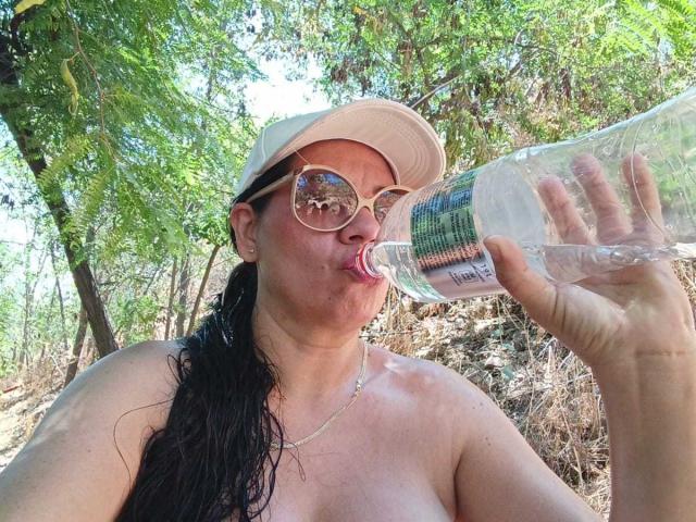 KinkyMaturee, a medium-built woman with fair skin, dark wet hair, and glasses, drinks from a clear plastic water bottle. She wears a beige cap and no shirt, exposing her upper chest. Her tongue is out, and she's looking up slightly. Her left hand holds the bottle. The background is a sunlit, wooded area with green leaves and dry underbrush. The photo is candid and casual, with natural light.